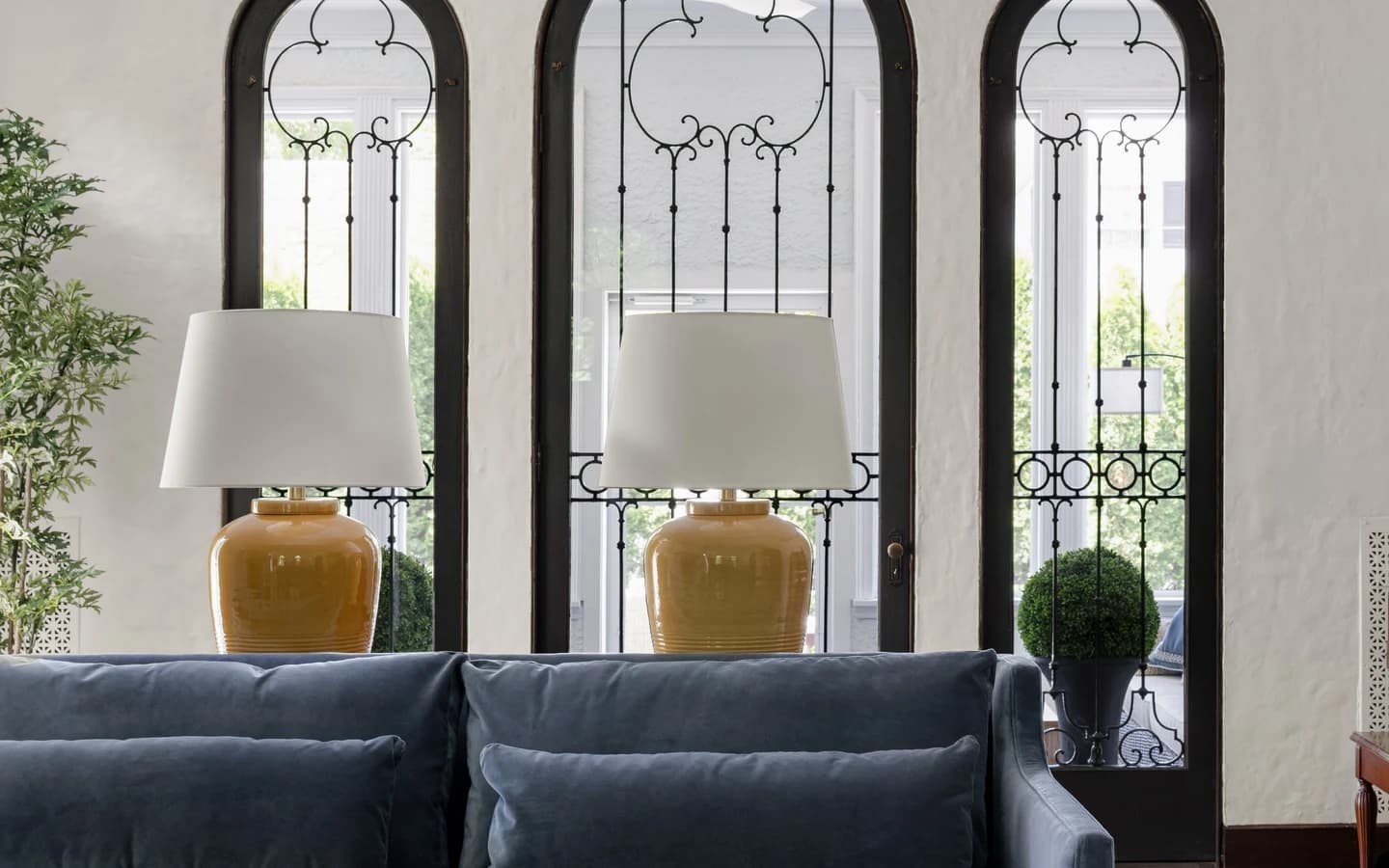 Blue velvet sofa with honey ceramic table lamps in front of arched windows with wrought iron detailing at a Blackstone Boulevard home in Providence, Rhode Island, designed by Hill & Harbor