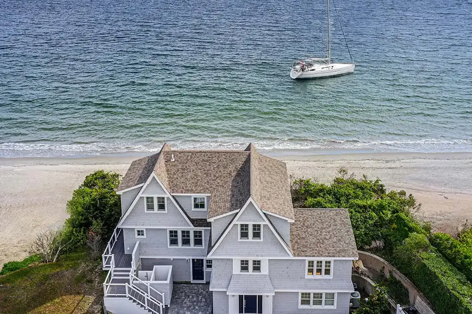 Aerial view of Project One Zero Eight, a grey shingle coastal home on the beach in Narragansett, Rhode Island, with a sailboat on the water beyond