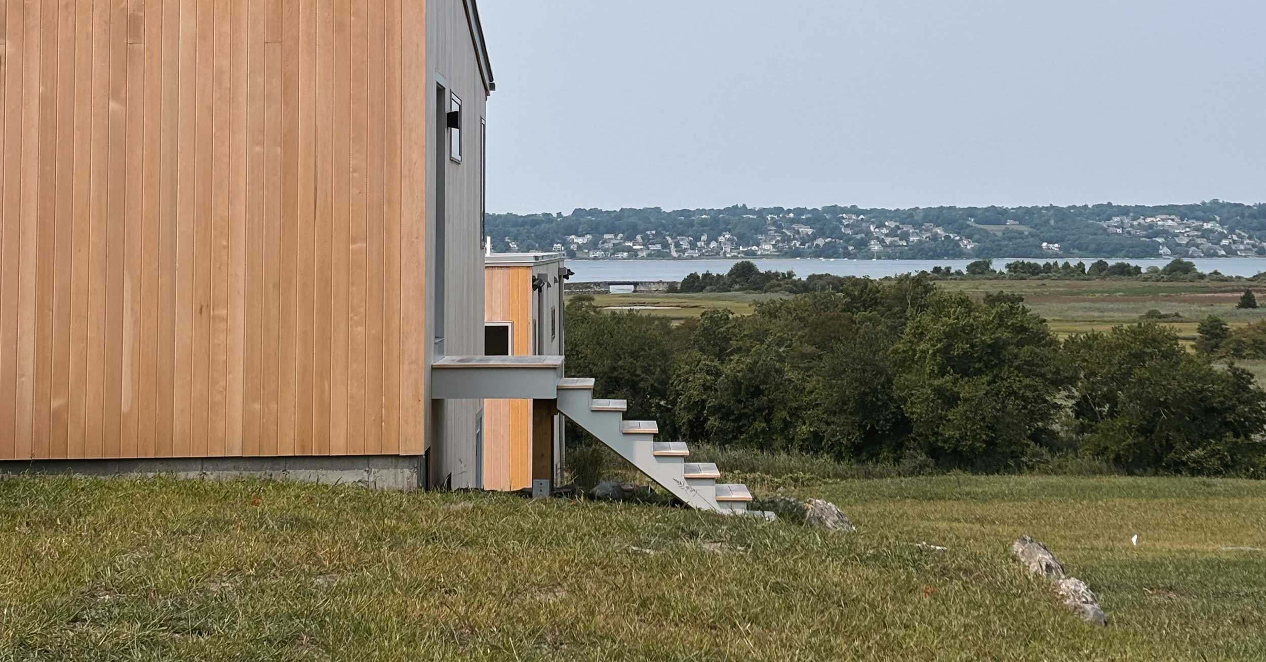 A modern coastal home in Tiverton features clean lines and natural wood siding. The new construction sits on a grassy hill overlooking a vast salt marsh and the Sakonnet River. Sleek exterior stairs lead to an elevated entrance, emphasizing an intentional connection to the Rhode Island landscape. The surrounding vista showcases lush greenery and distant waterfront properties under a soft, overcast sky.