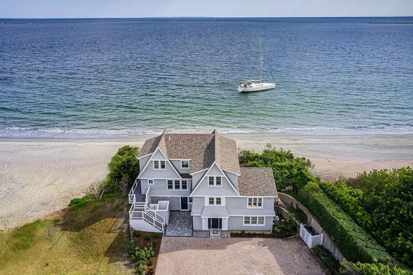 An aerial view of a coastal home renovation in Narragansett, Rhode Island, showcasing a multi-story, grey shingle house positioned directly on the beach with a sailboat anchored in the turquoise ocean water beyond.
