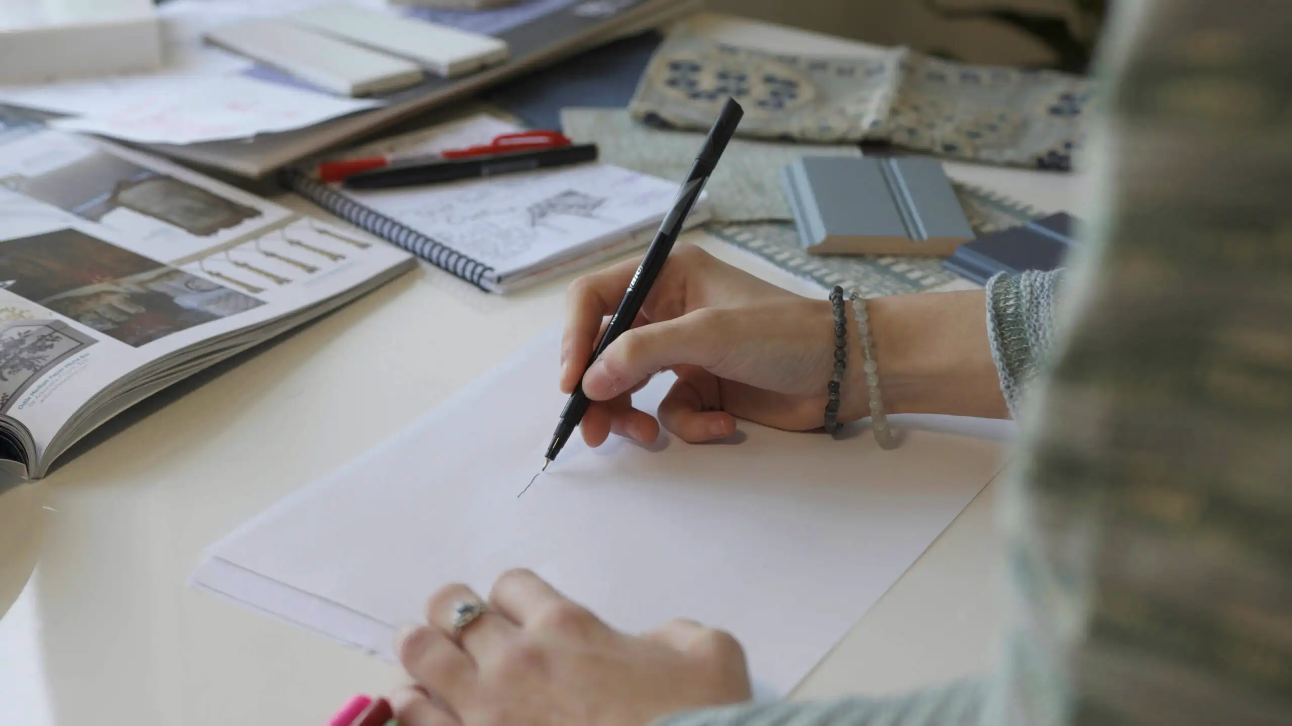 Close-up of hands sketching a line on blank paper with a black pen. An assortment of interior design materials surrounds the workspace, including patterned fabric swatches, painted wood trim samples, and an open design catalog. The person drawing wears beaded bracelets and a silver ring.