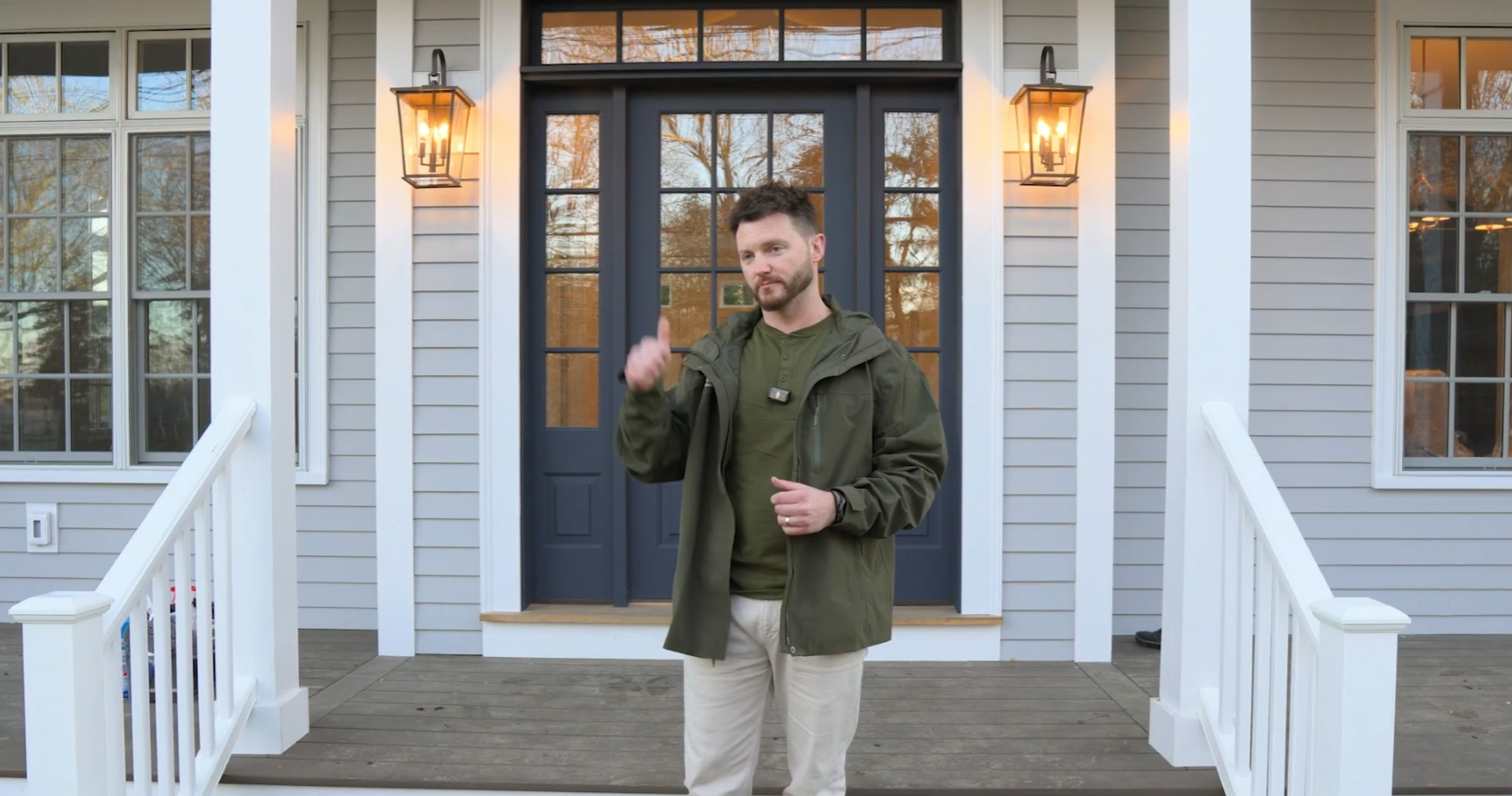 A man wearing an olive green jacket and a small lapel microphone stands on a wooden front porch. He gestures slightly while speaking. A dark blue glass-paneled front door, flanked by two lit wall lanterns, is directly behind him. The house features grey siding and white trim.