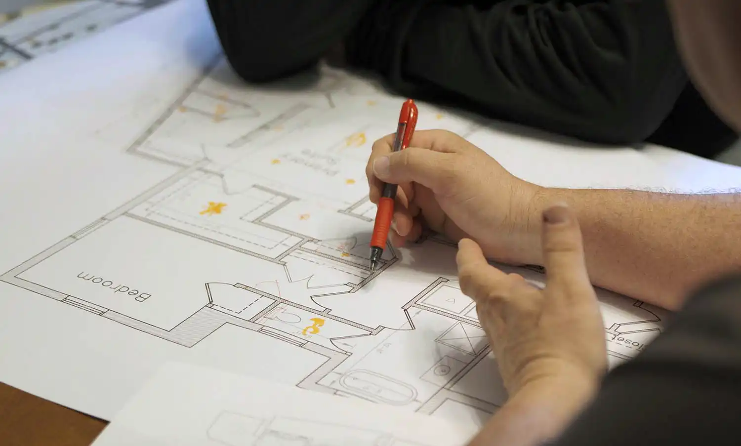 Close-up of hands holding a red mechanical pencil and pointing at a specific room on a printed architectural floor plan. Another person's arms rest on the table in the background as they review the blueprints together.